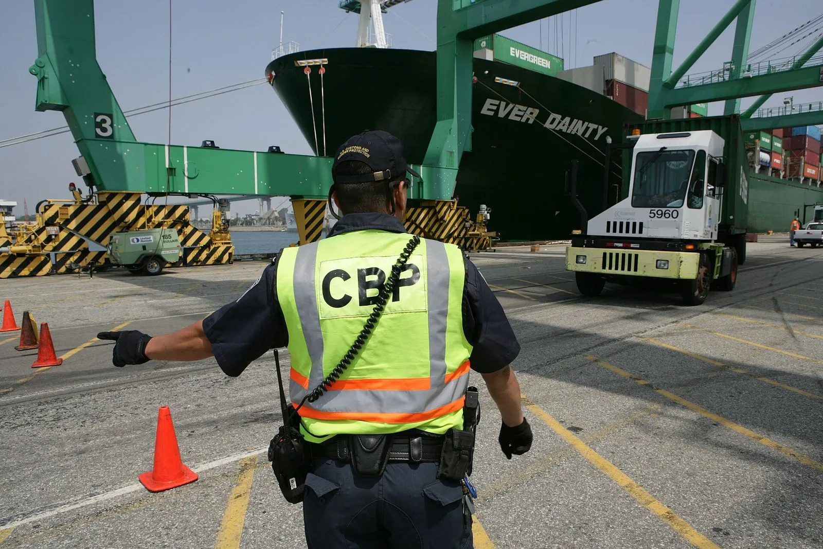 A Customs and Border Protection officer in a high-visibility vest directing a container truck inside a port terminal.