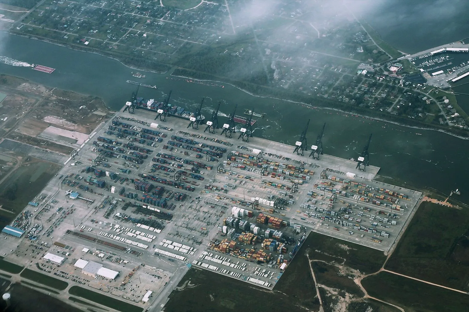 Aerial view of a riverfront container terminal with rows of stacked containers, gantry cranes and trucks on the wharf.