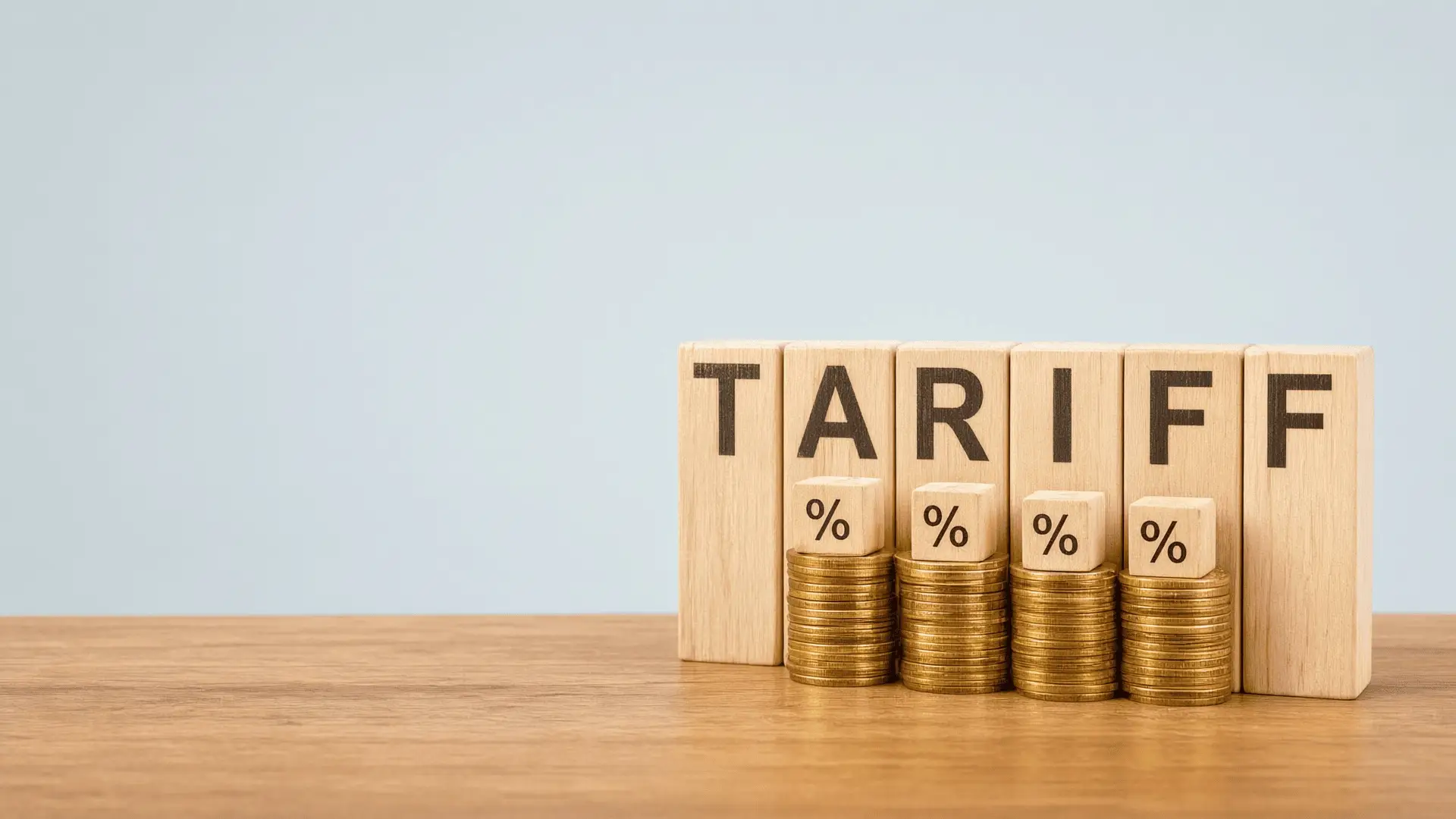 Bloques de madera que deletrean TARIFF junto a cuatro pilas de monedas de oro, cada una coronada por un bloque pequeño con un signo de porcentaje, sobre una mesa de madera